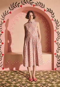 Rokav brez rožnate barve z rožnim potiskom, z ovratnikom in nabranim pasom. Model nosi ujemajoče rožnate balerinke in stoji na vzorčasti tlaščini.