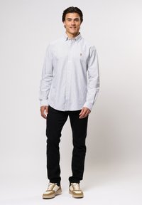 Cămașă în dungi, albastră și albă, cu guler clasic și logo pe piept. Asortată cu pantaloni negri și adidași crem.