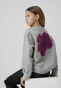 Felpa grigia con una grande stampa di un fiore di ibisco rosso scuro sulla schiena. Maniche a polsino e scollatura rotonda, abbinata a jeans scuri.