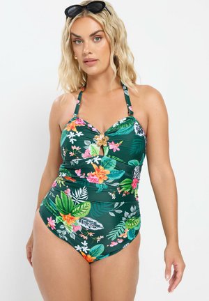 Lyshåret kvinde iført en grøn blomsterprintet badedragt med halterneck og en gulddetalje formet som en blomst samt sorte solbriller på hovedet.