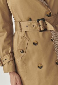 Beige trenčkot s dvojitými knoflíky, páskem v pase a kostičkovanou tkaninou na klopě. Texturovaný, voděodolný materiál.