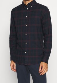 Man som bär en mörk marinblå skjorta med knappar, prydd med röda rutor och beige knappar, i kombination med mörk marinblå byxor.