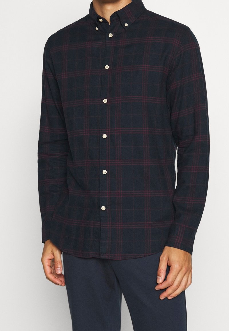 Man som bär en mörk marinblå skjorta med knappar, prydd med röda rutor och beige knappar, i kombination med mörk marinblå byxor.