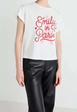 Weißes T-Shirt mit kurzen Ärmeln, rotem Text "Emily in Paris" und Eiffelturm-Grafik, getragen mit schwarzen Lederhosen an einer stehenden Person.