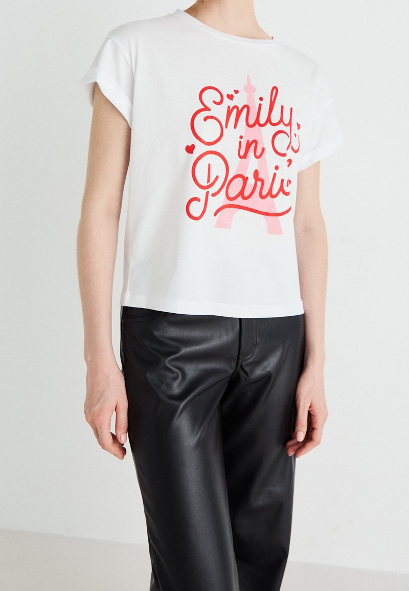 T-shirt blanc à manches courtes avec texte rouge "Emily in Paris" et graphique de la tour Eiffel, porté avec un pantalon en cuir noir sur une personne debout.