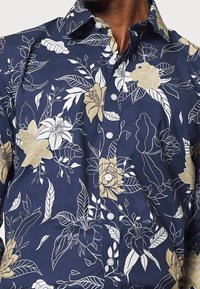 Camicia blu navy con grandi motivi floreali e foglie in bianco e beige, con chiusura frontale con bottoni e colletto standard. Materiale in cotone.