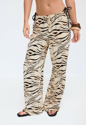 Pantalon ample à rayures tigre avec taille ajustable par cordon, porté avec des sandales noires et des bracelets épais sur une personne debout devant un fond uni.