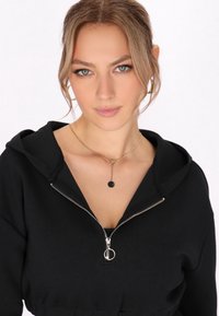 Felpa nera con zip e cappuccio, con una cerniera argentata e lacci. Indossata con una semplice collana con pendente rotondo. Tessuto dalla superficie liscia.