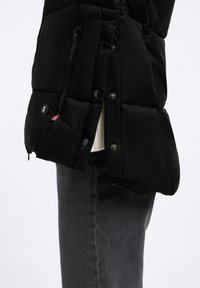 Czarna kurtka puchowa o pikowanej fakturze, z boczną kieszenią na zamek i zatrzaskami. Połączona z ciemnoszarymi dżinsami dla uzyskania warstwowego wyglądu.