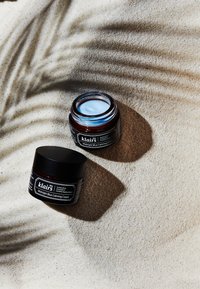 Klairs Midnight Blue Calming Cream krukker, mørk amberglass, sorte låg, med en lys blå krem synlig i den åpne krukken, på bakgrunn av sandaktig tekstur.