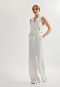 Weißes, gestreiftes, ärmelloses Oberteil mit Wickeldesign, kombiniert mit fließenden weißen Hosen. Flache, spitze Schuhe runden das Outfit ab.