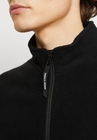 Svart fleecejacka med hög krage och dragkedjestängning. Har en svart dragkedja och en etikett med "JACK & JONES" i vit text.
