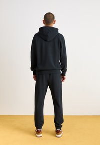 Sort hættetrøje og matchende joggingbukser lavet af blødt stof. Har ribbede manchetter, afslappet pasform og er sat sammen med sneakers.