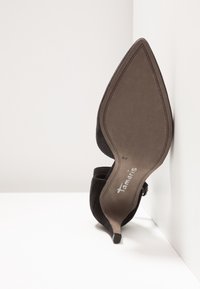 Une chaussure à talon haut noire à bout pointu reposant semelle contre un mur d'angle blanc, montrant la marque "Tamaris" et la taille 37.