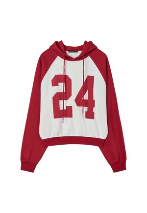 Jeune femme en tenue de sport rouge et blanche tenant une boîte à pizza, debout à côté d'une grande poutre métallique rouge sur un fond gris.
