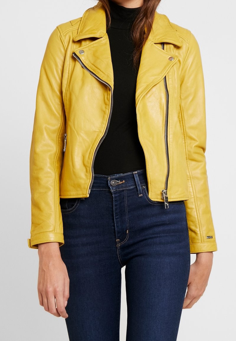Giubbotto in pelle gialla da biker con zip, colletto a piastra e vestibilità aderente. Indossato sopra un maglione nero a collo alto e abbinato a jeans blu.
