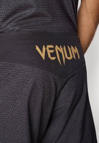 Calções desportivos pretos com um padrão texturizado, cintura lisa e um proeminente logótipo dourado "Venum" na parte de trás.