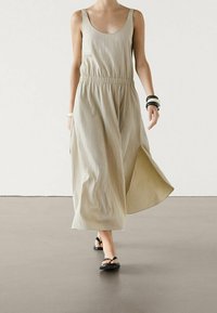 Robe sans manches beige clair avec une taille élastique resserrée, tissu fluide, fentes latérales, associée à des sandales plates noires et des bracelets audacieux.