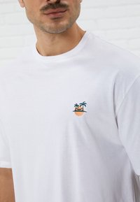 Weißes Baumwoll-T-Shirt mit einem kleinen gestickten Design eines Topfes mit Palmen und einem Gesicht. Einfacher Rundhalsausschnitt und kurze Ärmel.