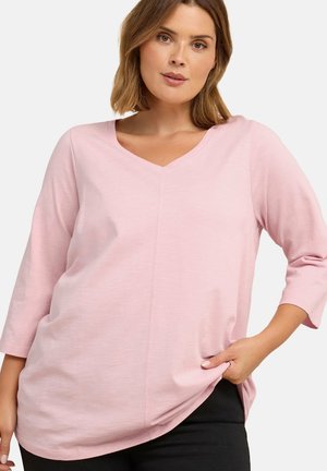 Kvinde iført lyserød, langærmet V-halsbluse med trekvartærmer, poserer med den ene hånd i lommen på sorte bukser på hvid baggrund.