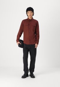 Camisa castanha de botão com dois bolsos no peito, jeans pretos, sapatos pretos, segurando uma mala preta; tecido suave e ajuste estruturado.