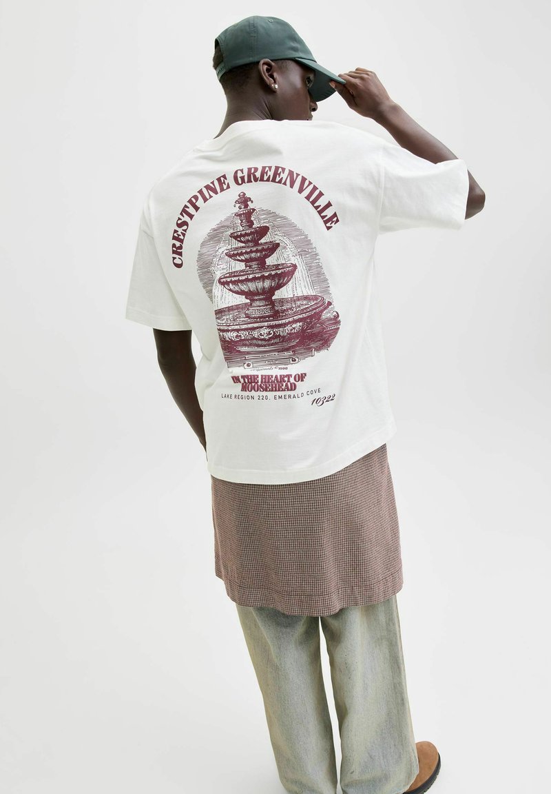 Osoba ubrana w biały t-shirt z dużą bordową grafiką fontanny i napisem, zieloną czapkę, kraciastą spódnicę na jasnych dżinsach oraz brązowe buty.