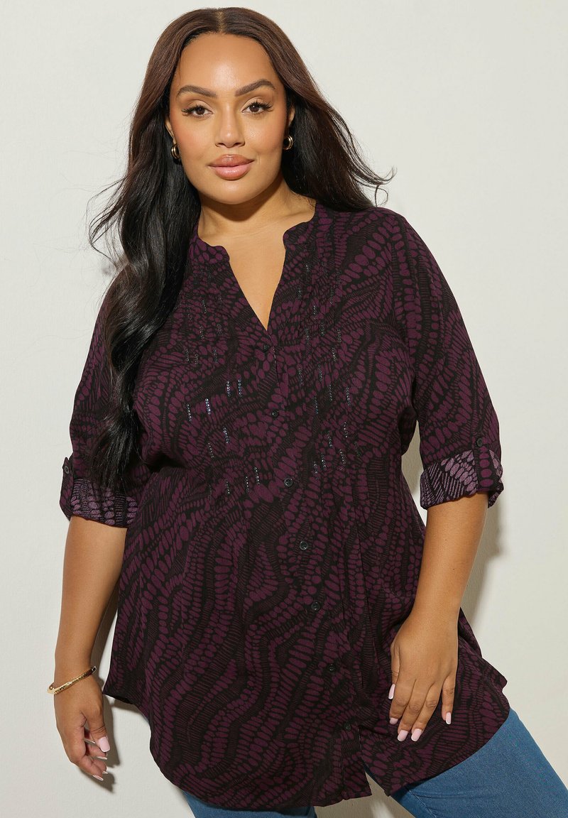 Una donna con lunghi capelli scuri indossa una camicia a maniche rigirate di colore viola a fantasia e jeans blu, su uno sfondo chiaro.