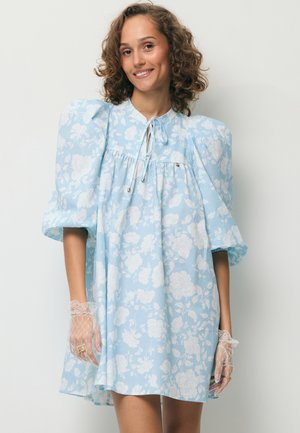 Vrouw met een lichtblauwe bloemenjurk met pofmouwen en kanten handschoenen, glimlachend tegen een effen lichte achtergrond.