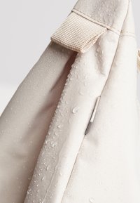 Coin d'un sac en tissu beige clair avec une sangle texturée, des gouttes d'eau sur la surface indiquant une résistance à l'eau ou un mouillage récent.