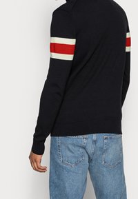 Stickad marinblå tröja med röda och vita randiga detaljer på ärmarna, med en ribbad krage och manschetter. Bärs med ljusblå jeans.