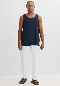 Top sin mangas en azul marino, de corte relajado, combinado con pantalones blancos holgados y sandalias marrones, con un diseño simple y una textura suave.