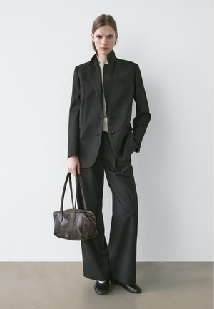 Femme portant un blazer noir cintré et un pantalon à jambes larges, tenant un sac à main en cuir sombre, se tenant devant un mur clair uni.