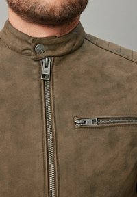 Chaqueta de cuero marrón con cuello alzado, cremallera frontal y bolsillo en el pecho. Presenta una textura suave y detalles de costura sutiles.
