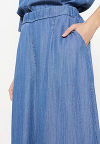 Denim krilo v svetlo modri barvi z elastičnim pasom in stranskimi žepi. Ima gladko teksturo in subtilne navpične črte.