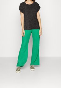 Top negro de manga corta y textura, combinado con pantalones de pierna ancha verde que presentan un dobladillo ondulado y un patrón a cuadros en los zapatos.