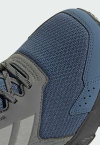 Sportssko med en struktureret blå mesh-overdel, grå syntetiske overlays, grå snørebånd og en sort gummisål med mønstrede detaljer.