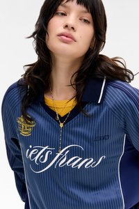 Camisa de malha riscada azul-marinho com um logótipo e letras brancas, apresentando um colarinho tradicional. Usada sobre uma camisa amarela, acentuada com colares em camadas.