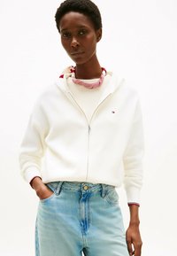Witte ritshoodie met rode en marineblauwe accenten, gedragen over een geribbelde witte top, gecombineerd met lichtblauwe high-waisted jeans.