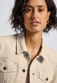 Femme avec des cheveux courts et bouclés portant des boucle d'oreilles en or et une veste beige à boutons, sur un fond gris clair.