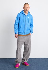 Sweat à capuche bleu à fermeture éclair avec une poche kangourou, associé à un pantalon en velours côtelé gris clair. Les chaussures sont roses et orange. Fond uni.