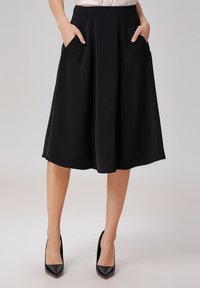 Jupe noire mi-longue, design évasé, tissu lisse, avec deux poches avant et une ceinture. Portée avec des talons aiguilles noirs.