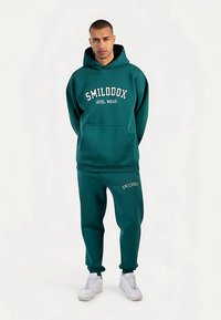 Turkusowa bluza z kapturem z białym nadrukiem "SMILODOX ATHL WEAR", z przednią kieszenią. Dopasowane spodnie dresowe, oba wykonane z miękkiego materiału, zestawione z białymi trampkami.