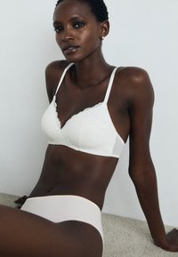 Hvit blonde bralette med bølgekant og myke stropper, kombinert med lys rosa sømløse undertøy, satt mot en nøytral bakgrunn.
