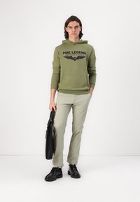 Zielona bluza z kapturem z napisem "PME Legend American Classic", kieszeń z przodu, zestawiona z jasnoniebieskimi spodniami i czarnymi butami, trzyma czarną torbę.