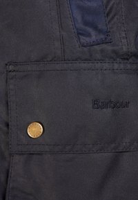 Donkere stof met een gladde textuur heeft een zak en een bronzen logo-knop. Zwarte geborduurde tekst leest "Barbour" op het oppervlak.