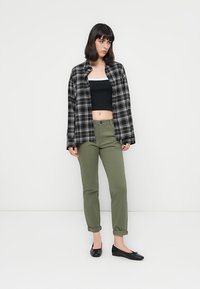 Kvinne med svart bob-frisyre iført svart og hvit rutet skjorte over svart crop-top, olivengrønne bukser og svarte ballerinaer, stående mot hvit vegg.