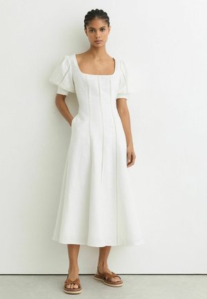 Moteris su kaspinu plaukuose stovi vilkėdama baltą midi suknelę su pūstomis rankovėmis ir kvadratiniu iškirpte, dėvi rudus plokščius sandalų.