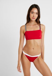 Punainen bikinitsetti, jossa on bandeau-toppi ohuilla mustilla hihnoilla ja matalavyötäröiset alushousut, joissa on valkoinen yksityiskohta. Sileä kangas, minimaalinen muotoilu.