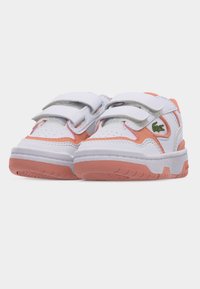 Λευκά και ροδακινί πάνινα παπούτσια για νήπια με δύο λευκές λουρίδες βέλκρο και πράσινο λογότυπο κροκόδειλου στο πλάι, εμφανιζόμενα από μπροστινή γωνία.