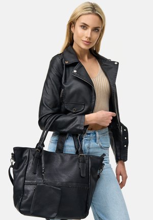 Zwarte leren handtas met dubbele handvaten, voorzien van een voorste ritszak en een getextureerde oppervlakte. Het model draagt een strakke zwarte jas en jeans.